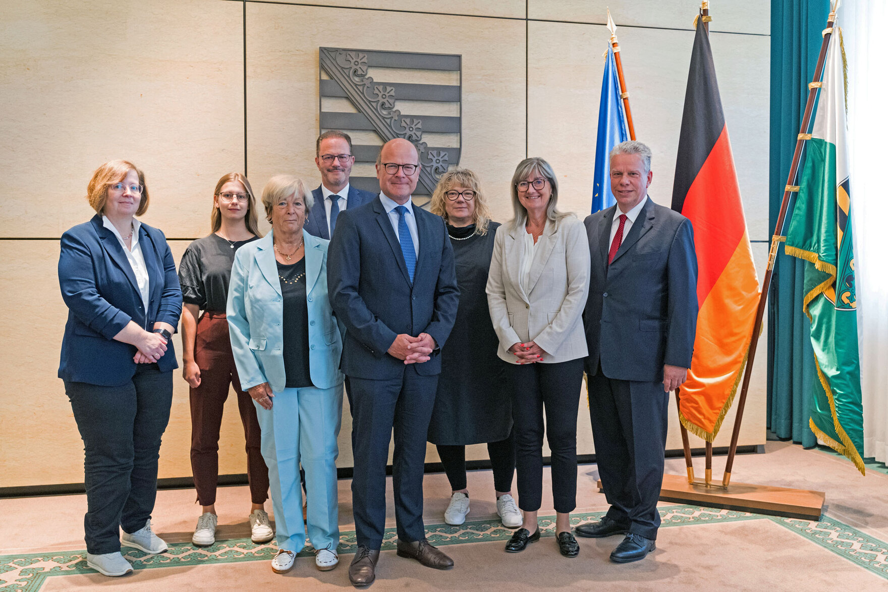 Männer und Fraune stehen vor einer Wand, an der das Wappen des Freistaates hängt.